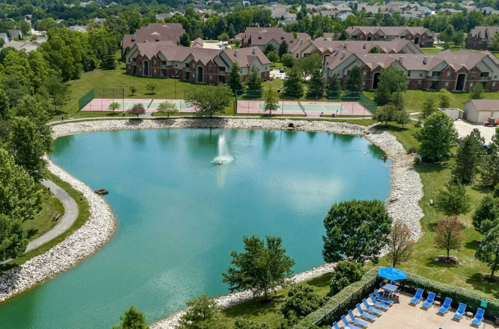 Aerial view of several trees, a pond with a walking path, 2 tennis courts, a poolside sundeck with chairs, and apartment buildings.