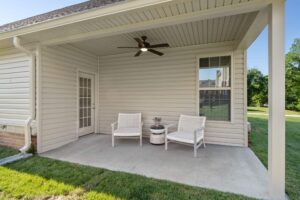 A large 3-bedroom cottage covered patio with a ceiling fan, 2 chairs, and a side table near a large green space with grass.